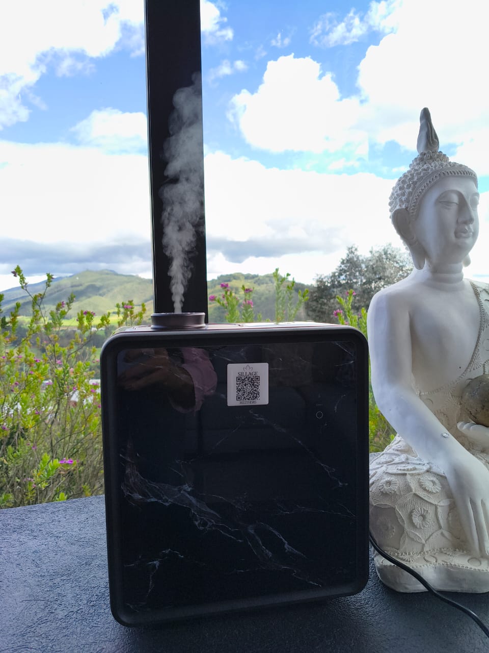 Difusor Sillage negro liberando una columna de vapor aromático junto a una figura de Buda blanca, con montañas al fondo.